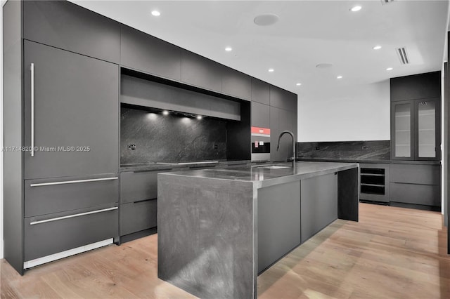 kitchen featuring light wood-type flooring, backsplash, beverage cooler, sink, and a large island with sink