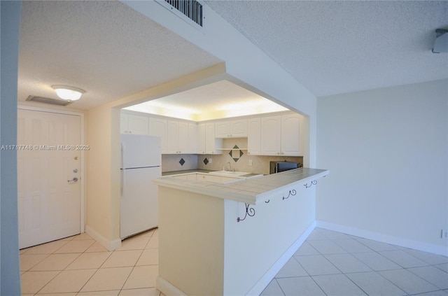 kitchen with visible vents, tile counters, white cabinets, freestanding refrigerator, and a peninsula