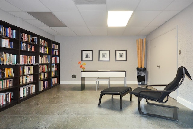 sitting room with a textured wall, wall of books, a paneled ceiling, and baseboards