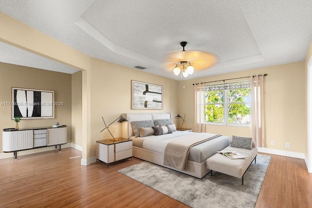 bedroom with a textured ceiling, hardwood / wood-style flooring, a raised ceiling, and ceiling fan