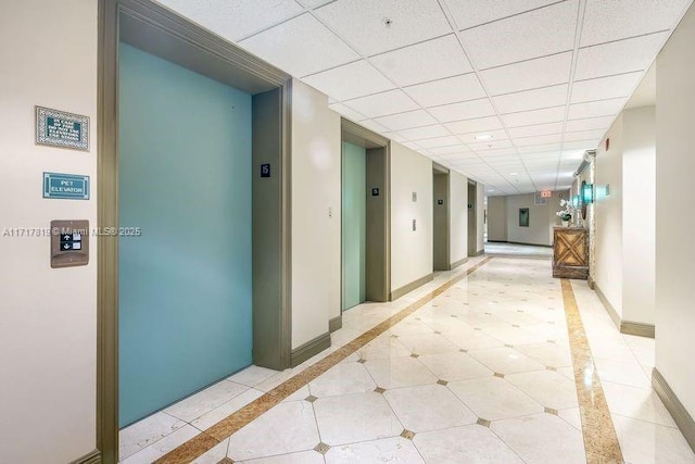 hallway with a drop ceiling, elevator, baseboards, and light tile patterned flooring
