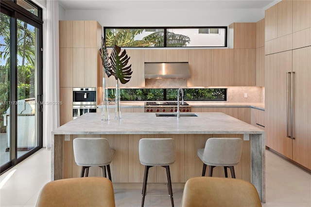 kitchen with a center island with sink, wall chimney exhaust hood, light brown cabinets, and sink