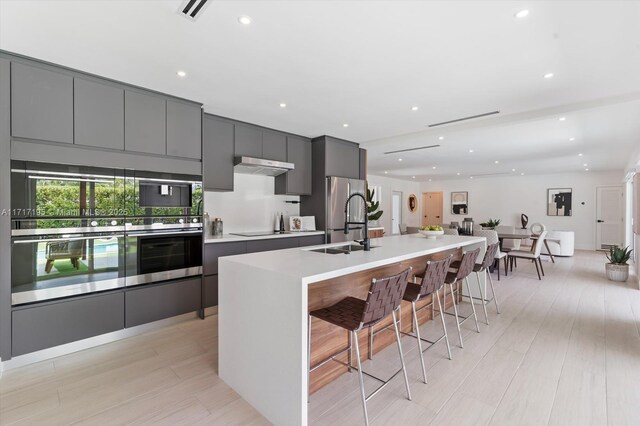 kitchen featuring a kitchen bar, gray cabinetry, an island with sink, stainless steel appliances, and exhaust hood