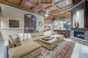 living room with coffered ceiling, ornamental molding, a stone fireplace, ceiling fan, and beamed ceiling