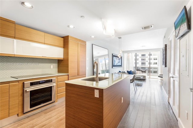 kitchen with tasteful backsplash, black electric cooktop, sink, oven, and an island with sink