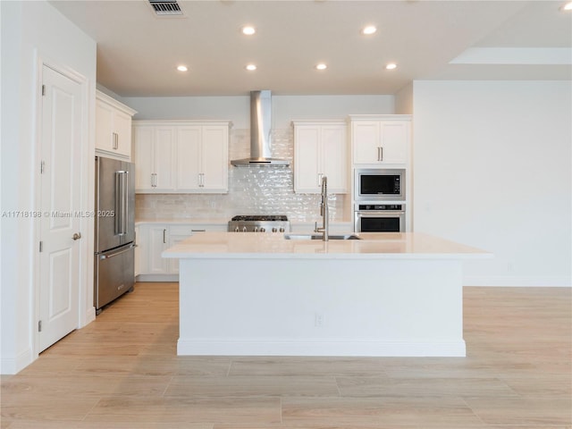 kitchen featuring stainless steel appliances, a kitchen island with sink, sink, wall chimney range hood, and white cabinets