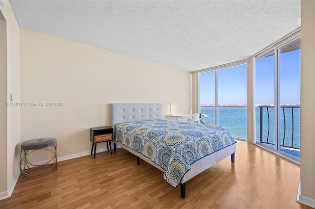 bedroom with access to outside, expansive windows, a water view, a textured ceiling, and wood-type flooring