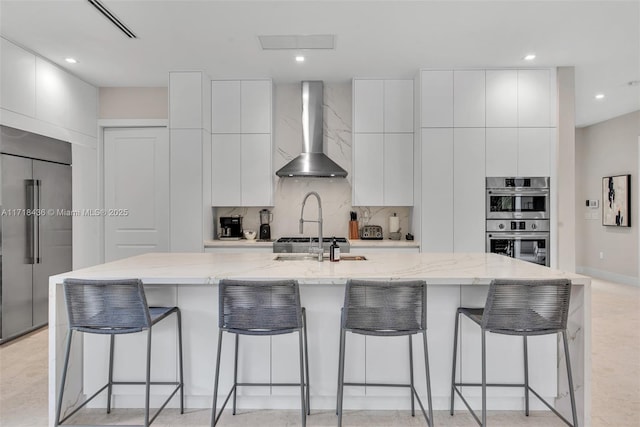 kitchen featuring sink, wall chimney exhaust hood, stainless steel appliances, a kitchen island with sink, and white cabinets