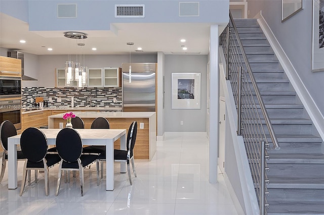 kitchen with wall chimney exhaust hood, built in appliances, decorative light fixtures, light tile patterned floors, and backsplash