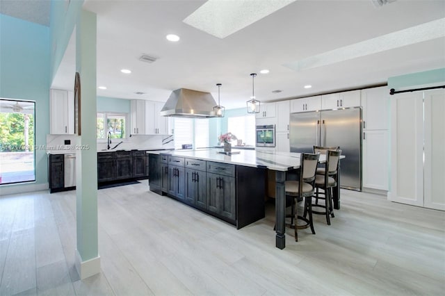 kitchen with stainless steel appliances, white cabinetry, hanging light fixtures, and wall chimney exhaust hood