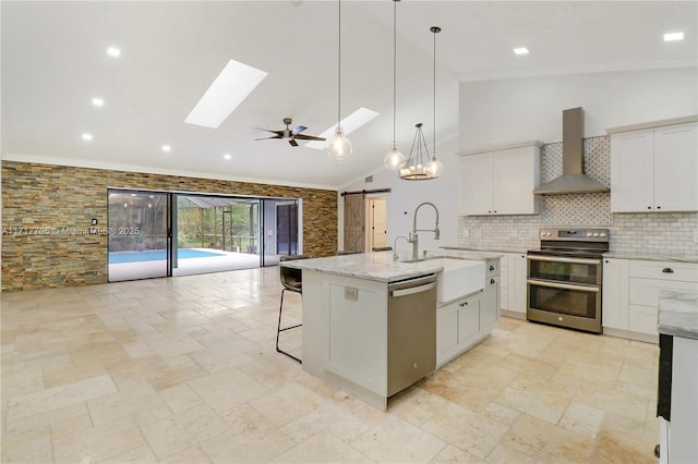 kitchen with a skylight, appliances with stainless steel finishes, white cabinetry, a barn door, and wall chimney exhaust hood