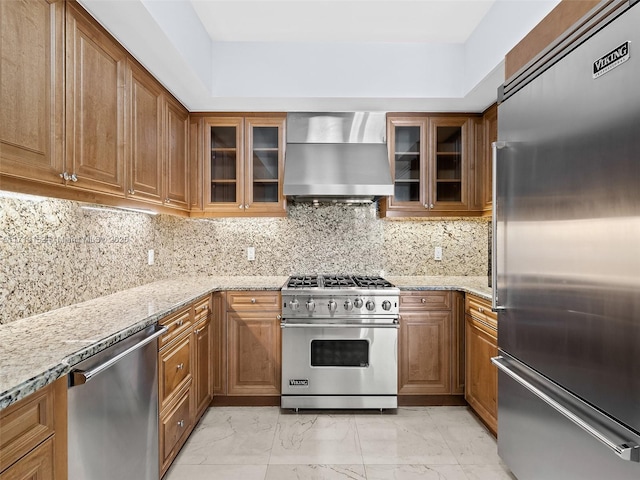 kitchen with light stone countertops, high end appliances, wall chimney range hood, and tasteful backsplash