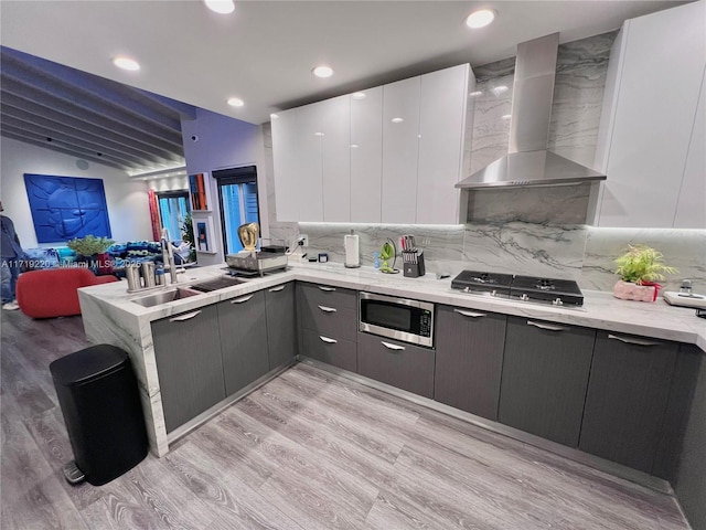 kitchen featuring wall chimney range hood, decorative backsplash, light wood-type flooring, appliances with stainless steel finishes, and white cabinetry