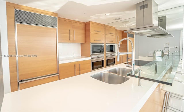 kitchen featuring island exhaust hood, built in appliances, light brown cabinets, and sink