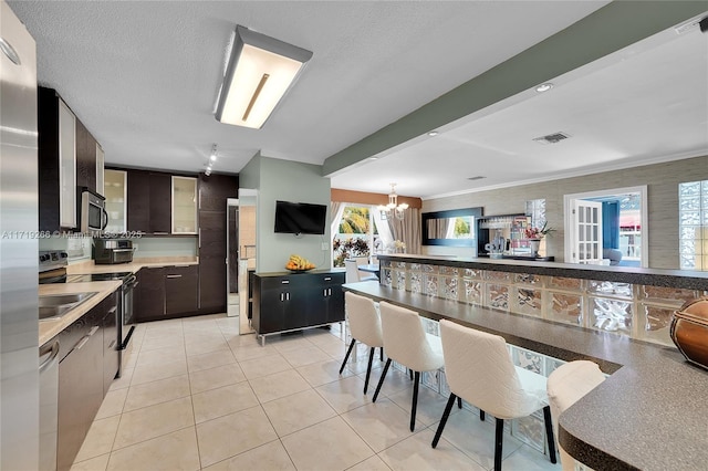 kitchen featuring dark brown cabinetry, an inviting chandelier, appliances with stainless steel finishes, light tile patterned floors, and ornamental molding