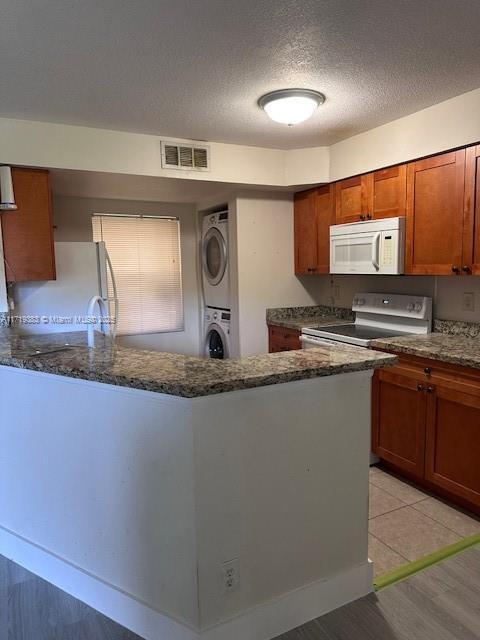 kitchen with a textured ceiling, white appliances, stacked washer / dryer, and dark stone counters