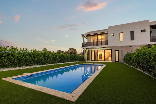 back house at dusk with a fenced in pool, a balcony, and a lawn