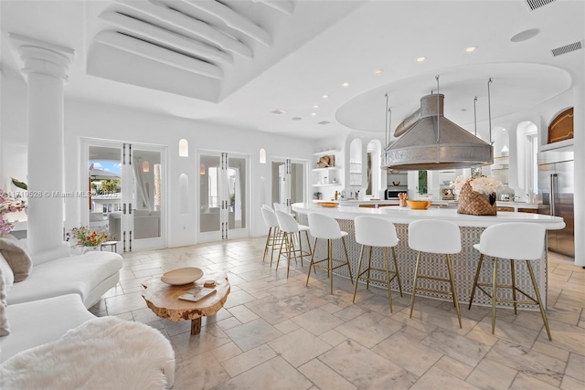 kitchen featuring pendant lighting, stainless steel built in fridge, ornate columns, island range hood, and a kitchen bar