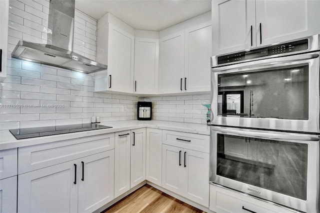 kitchen featuring stainless steel double oven, wall chimney range hood, white cabinets, and black electric cooktop