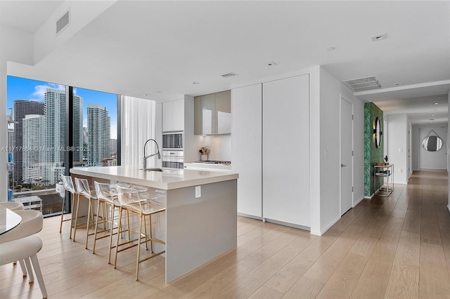 kitchen with oven, light hardwood / wood-style floors, a center island with sink, a breakfast bar, and white cabinetry