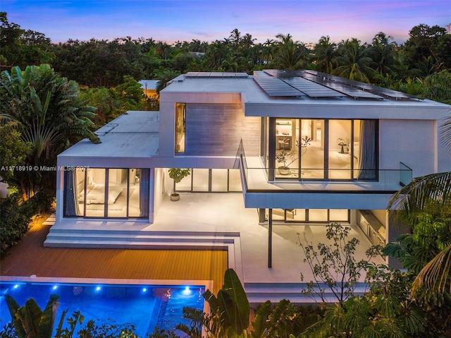 back house at dusk featuring solar panels, a patio area, a pool, and a balcony