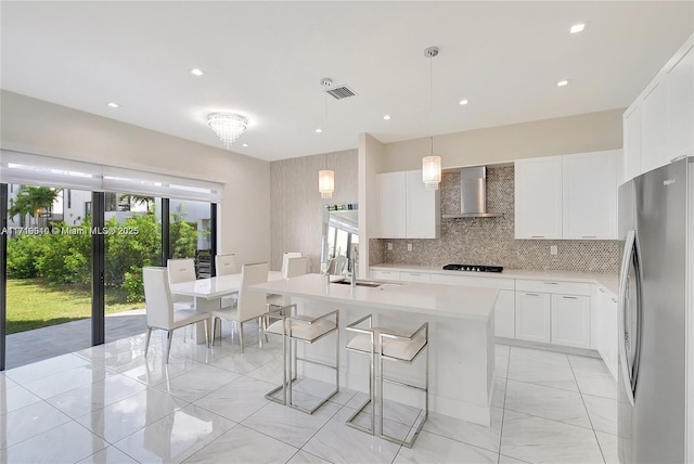 kitchen featuring wall chimney exhaust hood, a center island with sink, white cabinetry, hanging light fixtures, and stainless steel refrigerator