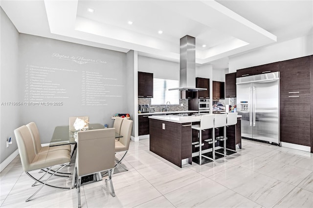 kitchen with a kitchen bar, stainless steel built in refrigerator, island range hood, a raised ceiling, and a kitchen island