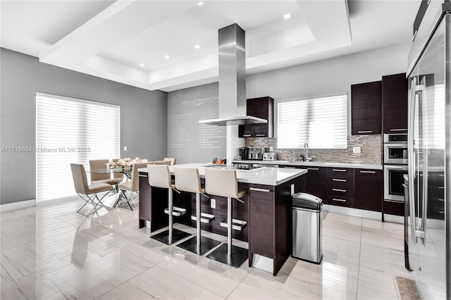 kitchen with a tray ceiling, island exhaust hood, sink, and a kitchen island