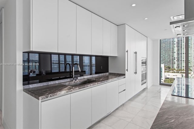 kitchen featuring white cabinetry, sink, stainless steel oven, a wall of windows, and dark stone counters