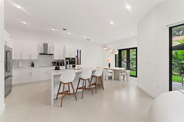 kitchen with white cabinets, stainless steel fridge, a center island with sink, and wall chimney exhaust hood