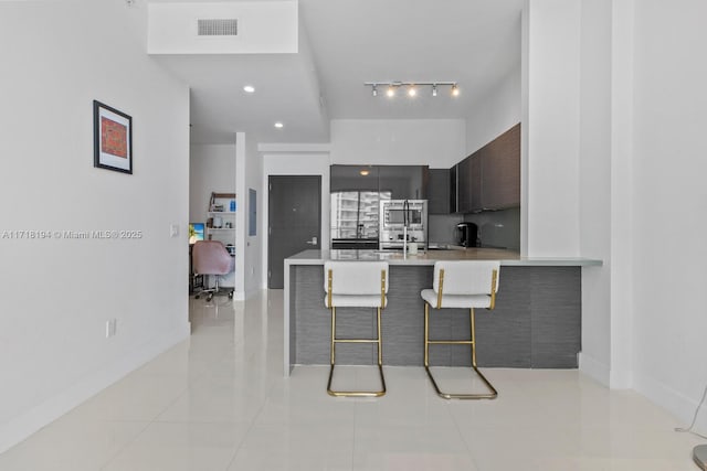 kitchen with stainless steel microwave, kitchen peninsula, a breakfast bar, light tile patterned flooring, and dark brown cabinets