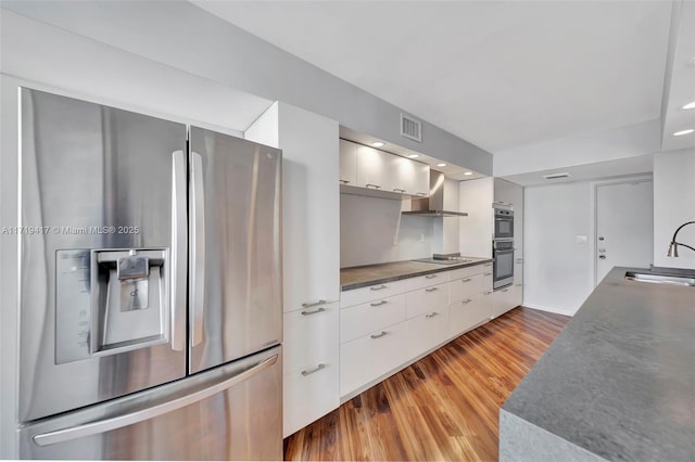 kitchen featuring stainless steel appliances, wall chimney range hood, sink, white cabinets, and light hardwood / wood-style floors