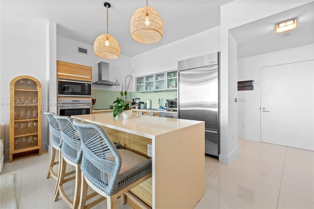 kitchen with a center island, wall chimney range hood, built in appliances, pendant lighting, and a breakfast bar