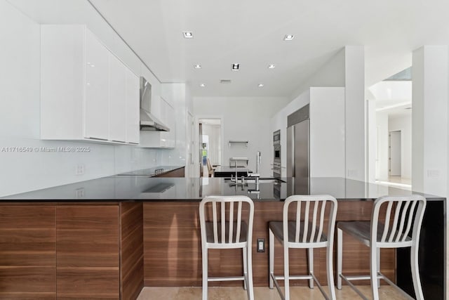 kitchen with built in appliances, kitchen peninsula, wall chimney range hood, white cabinets, and a kitchen breakfast bar