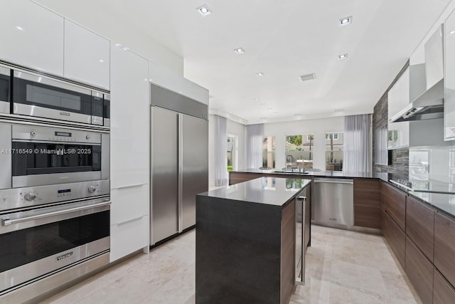 kitchen featuring built in appliances, wall chimney exhaust hood, a breakfast bar, a kitchen island, and white cabinetry