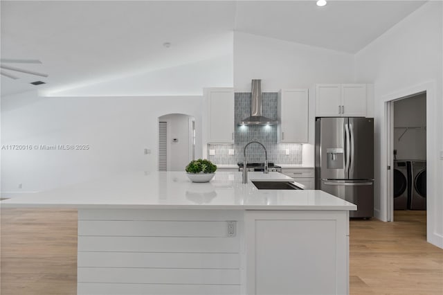 kitchen with vaulted ceiling, wall chimney range hood, washing machine and clothes dryer, white cabinets, and stainless steel fridge with ice dispenser