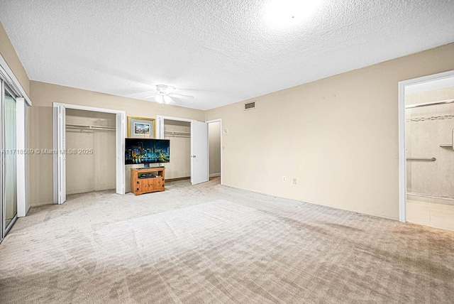 unfurnished bedroom featuring ensuite bath, a textured ceiling, two closets, light colored carpet, and ceiling fan