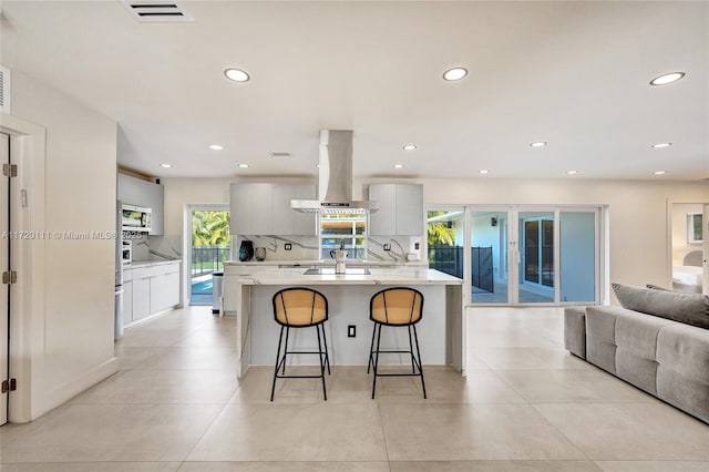 kitchen featuring decorative backsplash, island exhaust hood, white cabinetry, a breakfast bar area, and an island with sink