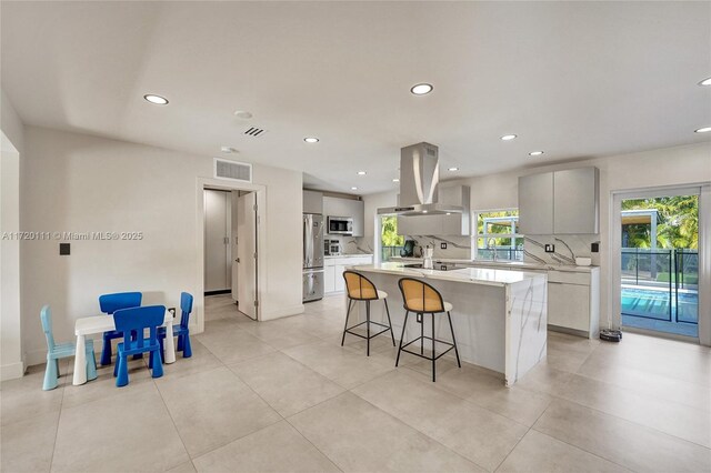 kitchen featuring stainless steel appliances, backsplash, island range hood, a breakfast bar, and a center island with sink