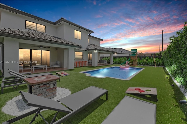 pool at dusk with a lawn, ceiling fan, a playground, and a patio