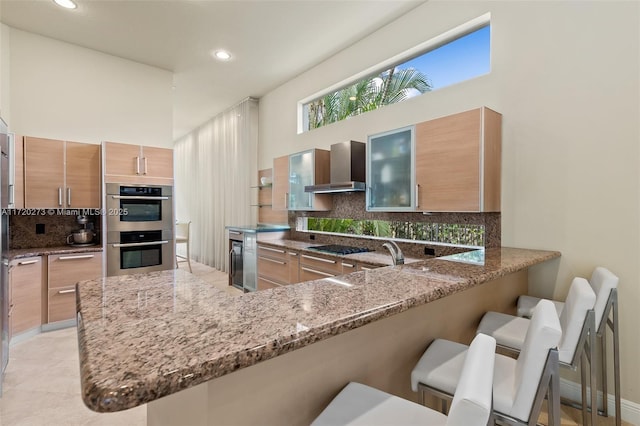 kitchen with kitchen peninsula, a kitchen bar, stainless steel double oven, and wall chimney exhaust hood