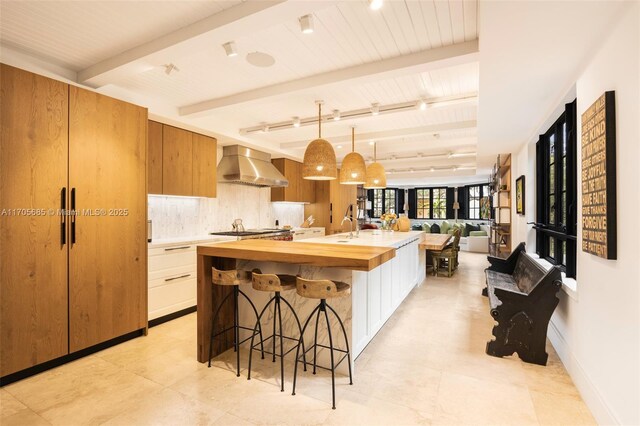 kitchen with white cabinetry, wall chimney exhaust hood, backsplash, decorative light fixtures, and a kitchen island with sink