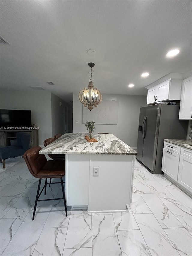 kitchen featuring stainless steel refrigerator with ice dispenser, light stone countertops, decorative light fixtures, a kitchen island, and white cabinetry