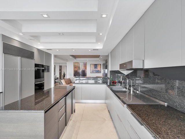 kitchen with a raised ceiling, sink, dark stone countertops, light tile patterned floors, and white cabinetry