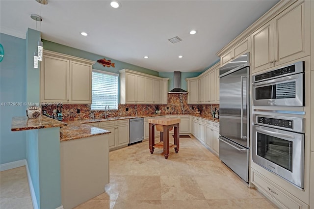 kitchen featuring wall chimney exhaust hood, a peninsula, cream cabinets, stainless steel appliances, and backsplash