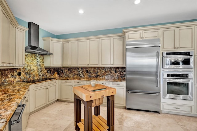 kitchen featuring backsplash, stone counters, cream cabinets, wall chimney range hood, and stainless steel appliances