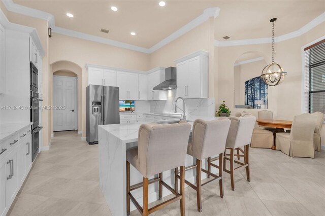 kitchen with light stone countertops, appliances with stainless steel finishes, wall chimney exhaust hood, decorative light fixtures, and white cabinetry