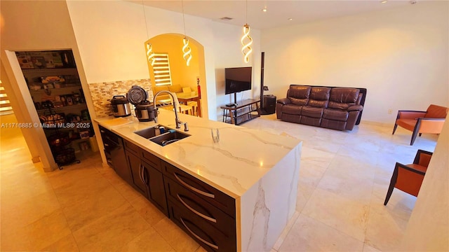 kitchen featuring dark brown cabinetry, light stone counters, open floor plan, pendant lighting, and a sink