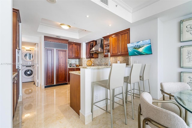 kitchen featuring a tray ceiling, paneled built in fridge, a peninsula, wall chimney range hood, and stacked washer / drying machine