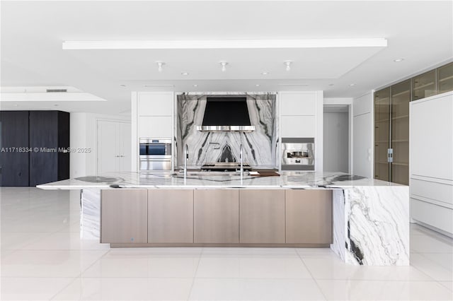 kitchen featuring wall chimney exhaust hood, light stone countertops, white cabinets, and a spacious island